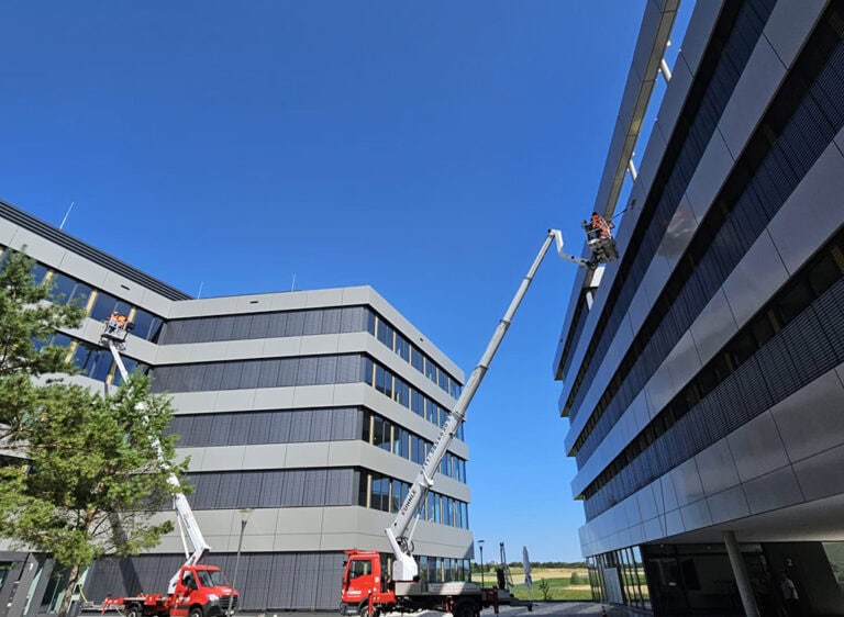 Zwei Personen auf Hebebühnen arbeiten an modernen grauen Bürogebäuden unter blauem Himmel.