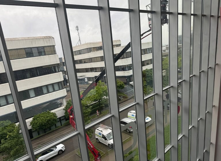 Blick durch ein Fenster auf Gebäude im Regen mit parkenden Fahrzeugen und Bäumen.