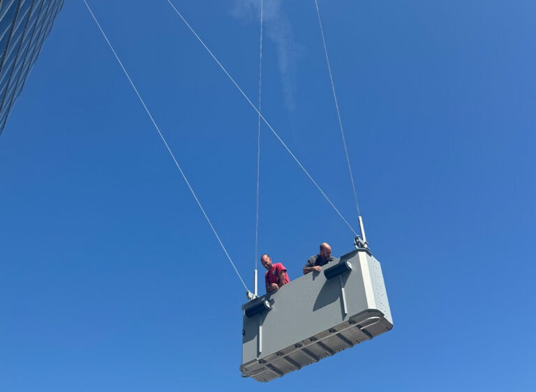 Zwei Männer in Sicherheitswesten schauen lächelnd aus einer schwebenden Plattform vor blauem Himmel.