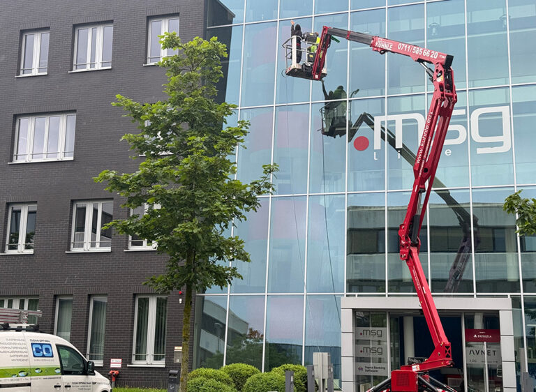 Ein Bauarbeiter auf einem roten Hebearm reinigt Fenster eines modernen Bürogebäudes mit Glasfassade.