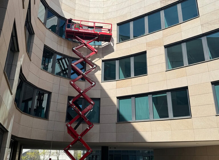 Ein Arbeiter in einem roten Hebebühnenlift vor einem modernen, beigen Bürogebäude.