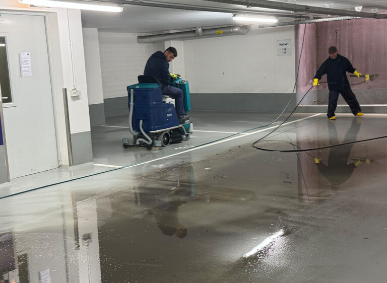 Zwei Männer reinigen das Parkhaus. Einer fährt eine Putzmaschine, der andere hält einen Schlauch.